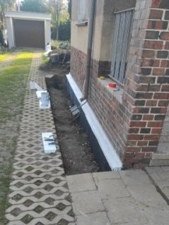 Odkopany fundament budynku z izolacją pionową, widoczne bloczki betonowe i kostka brukowa na zewnątrz. Prace ziemne w toku, przygotowanie do dalszych etapów izolacji.