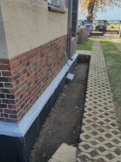 Odkopane fundamenty domu z widoczną izolacją pionową, ceglaną elewacją i chodnikiem z ażurowych płyt. Prace ziemne w toku, widoczne rury spustowe i fragment trawnika.