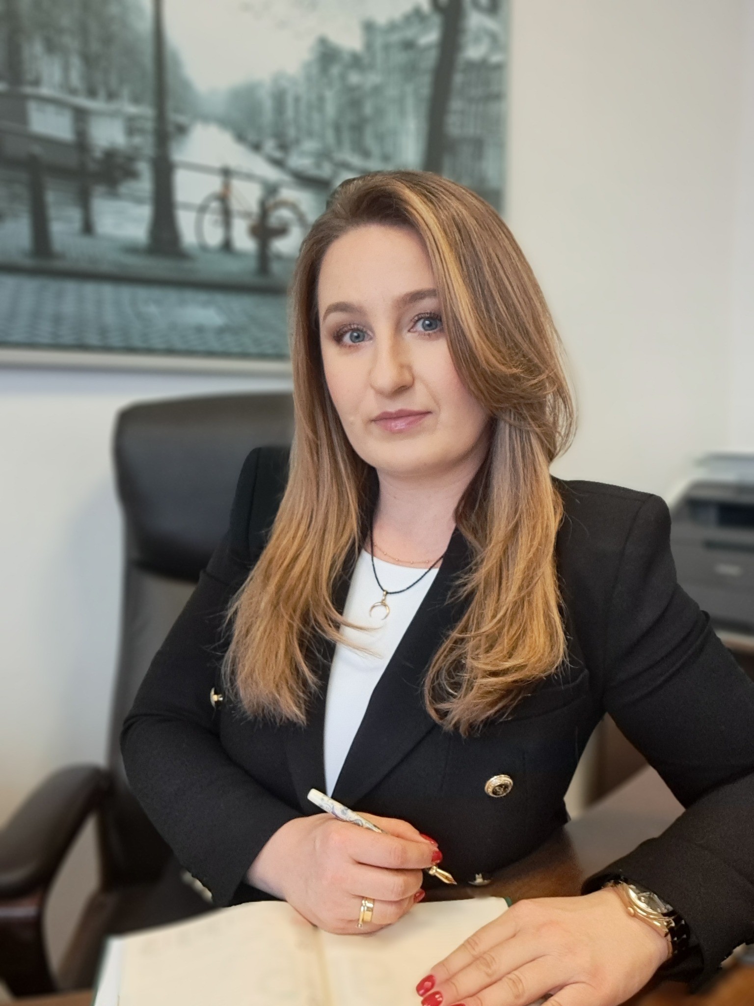 Profesjonalny portret kobiety w czarnej marynarce, siedzącej za biurkiem z notatnikiem i długopisem, w tle obraz z motywem miejskim. Ujęcie z bliska, naturalne światło.