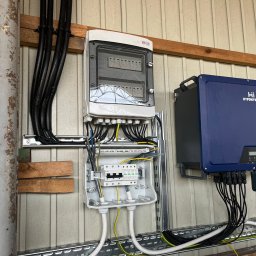 PHX SPÓŁKA Z OGRANICZONĄ ODPOWIEDZIALNOŚCIĄ - Rozdzielnica elektryczna z zabezpieczeniami przepięciowymi i falownik HYPONTECH zamontowane na ścianie z blachy trapezowej, widoczne przewody w peszlach i korytkach kablowych.