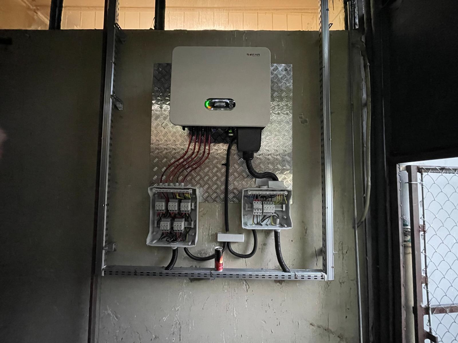 Zamontowany inwerter solarny marki SOFAR na aluminiowej płycie, z podłączonymi przewodami i dwiema skrzynkami elektrycznymi na szarej ścianie w pomieszczeniu gospodarczym.