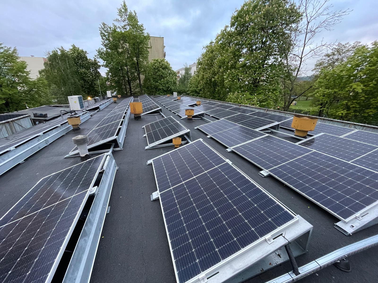 Panele słoneczne na płaskim dachu budynku, widok z góry, z kominami wentylacyjnymi i drzewami w tle.