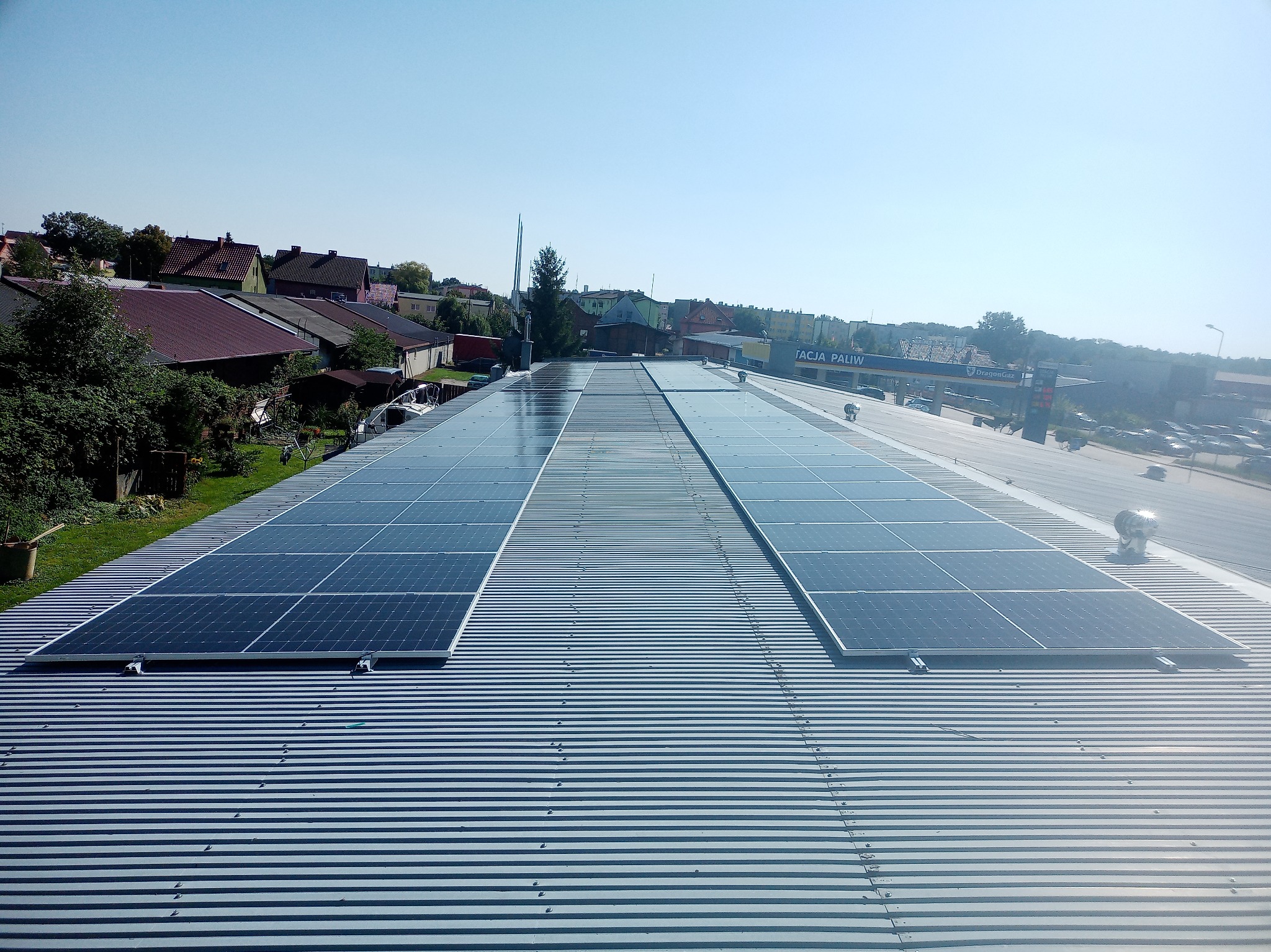 Instalacja fotowoltaiczna na dachu budynku przemysłowego, widok z góry, w tle zabudowa mieszkalna i stacja paliw.