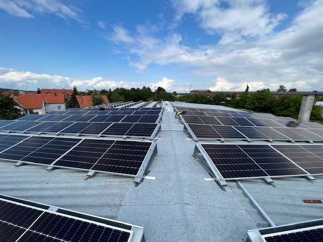 Montaż paneli słonecznych na dachu budynku przemysłowego, widok z góry, pracownik w odblaskowej kamizelce na dachu, błękitne niebo z chmurami w tle.