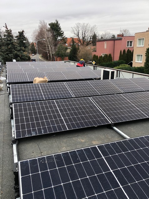 Montaż paneli słonecznych na płaskim dachu budynku mieszkalnego, widoczny pracownik w żółtym kasku i torba z narzędziami, w tle zabudowa miejska.