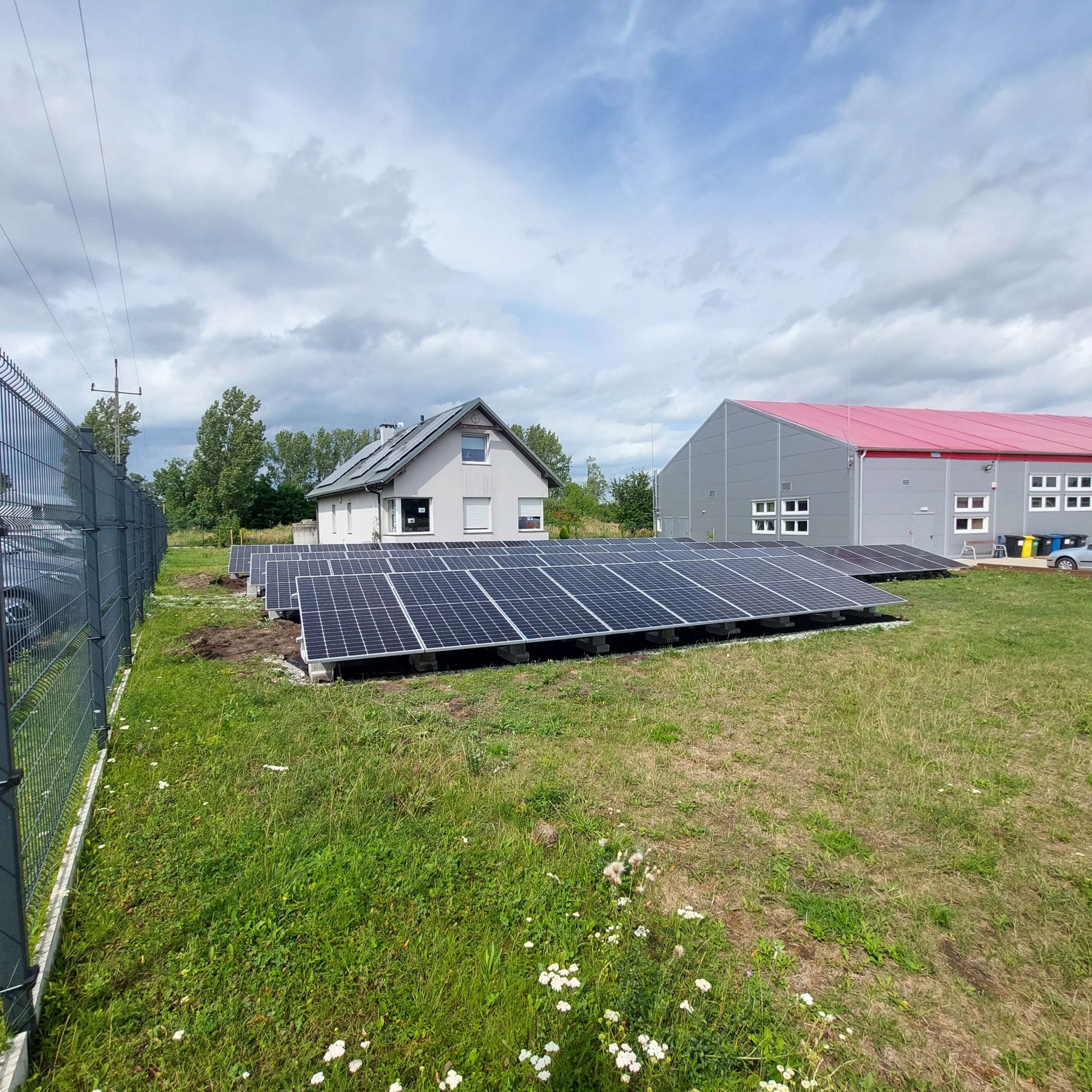 Instalacja paneli słonecznych na trawniku przed domem jednorodzinnym i budynkiem przemysłowym, widoczne kable i elementy montażowe, pochmurne niebo w tle.