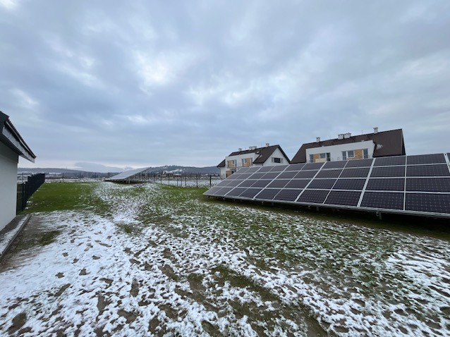 Rozległa instalacja paneli słonecznych na trawiastym terenie z resztkami śniegu, w tle budynki mieszkalne pod pochmurnym niebem.