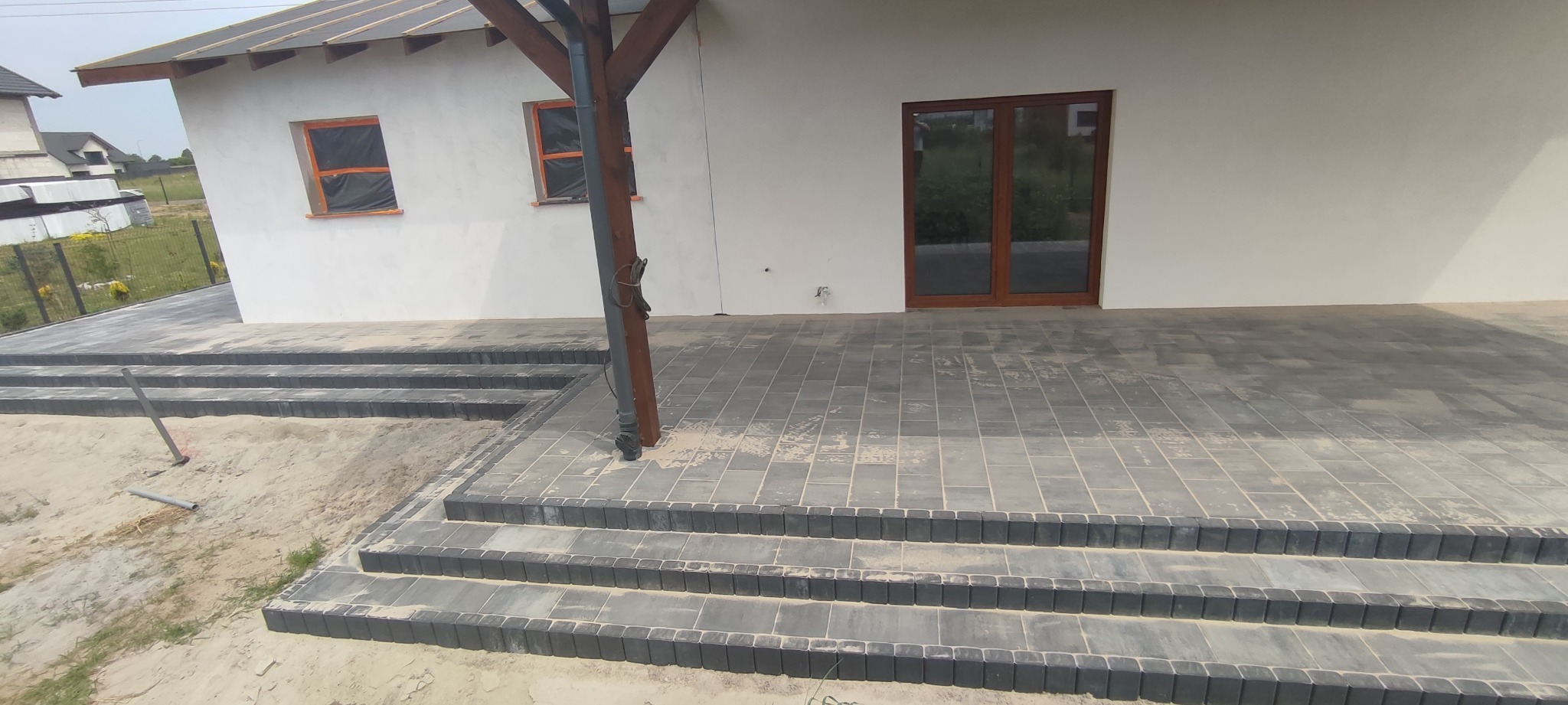 Nowo ułożony taras z szarej kostki brukowej prowadzący do domu z drewnianymi oknami i drzwiami tarasowymi, widoczne schody z kostki i piasek po bokach.