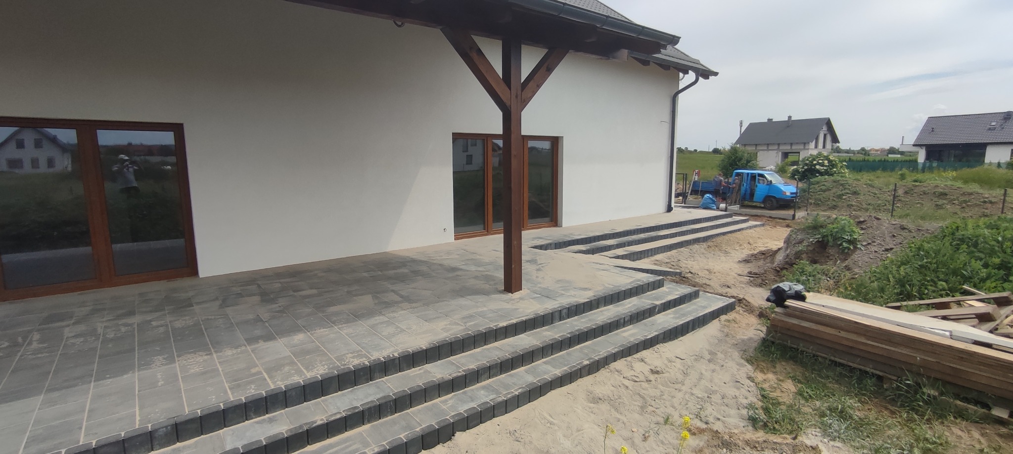 Nowoczesny taras z szarej kostki brukowej i schodami w Gdańsku, zadaszony drewnianą konstrukcją, w tle widoczny jasny budynek i ekipa budowlana przy niebieskim samochodzie dostawczym.