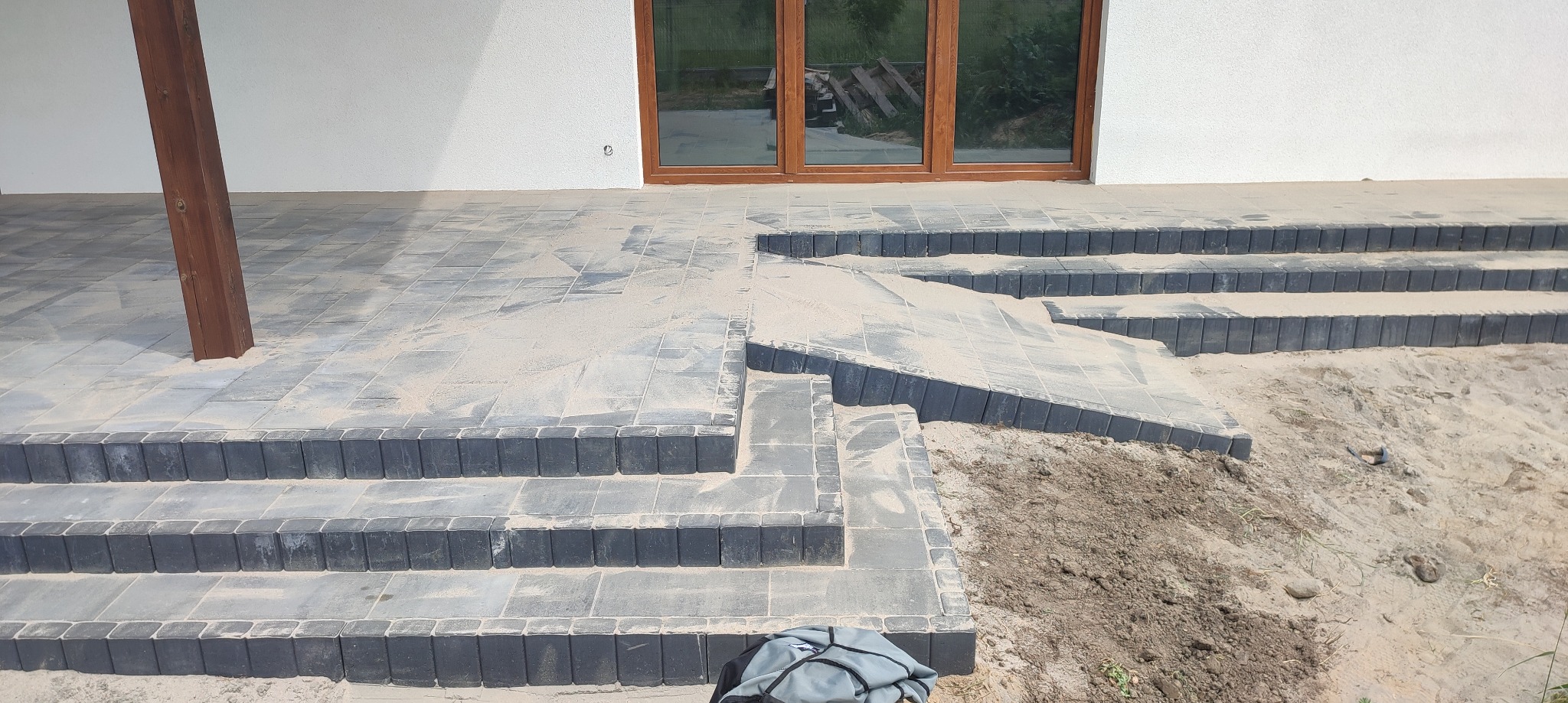 Schody wejściowe do domu wyłożone szarą kostką brukową, zasypane piaskiem, z widocznym fragmentem trawnika i workiem roboczym w rogu.