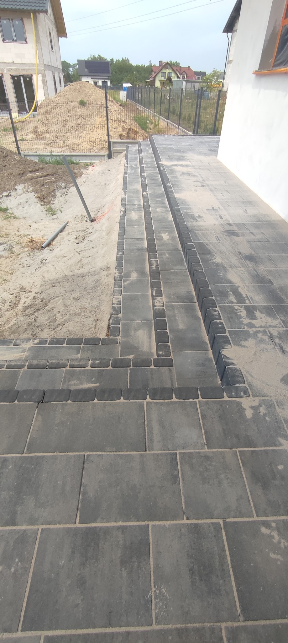Ułożona, szara kostka brukowa tworząca ścieżkę z odpływem liniowym, widoczny fragment posesji w trakcie prac wykończeniowych, z hałdą piasku w tle.