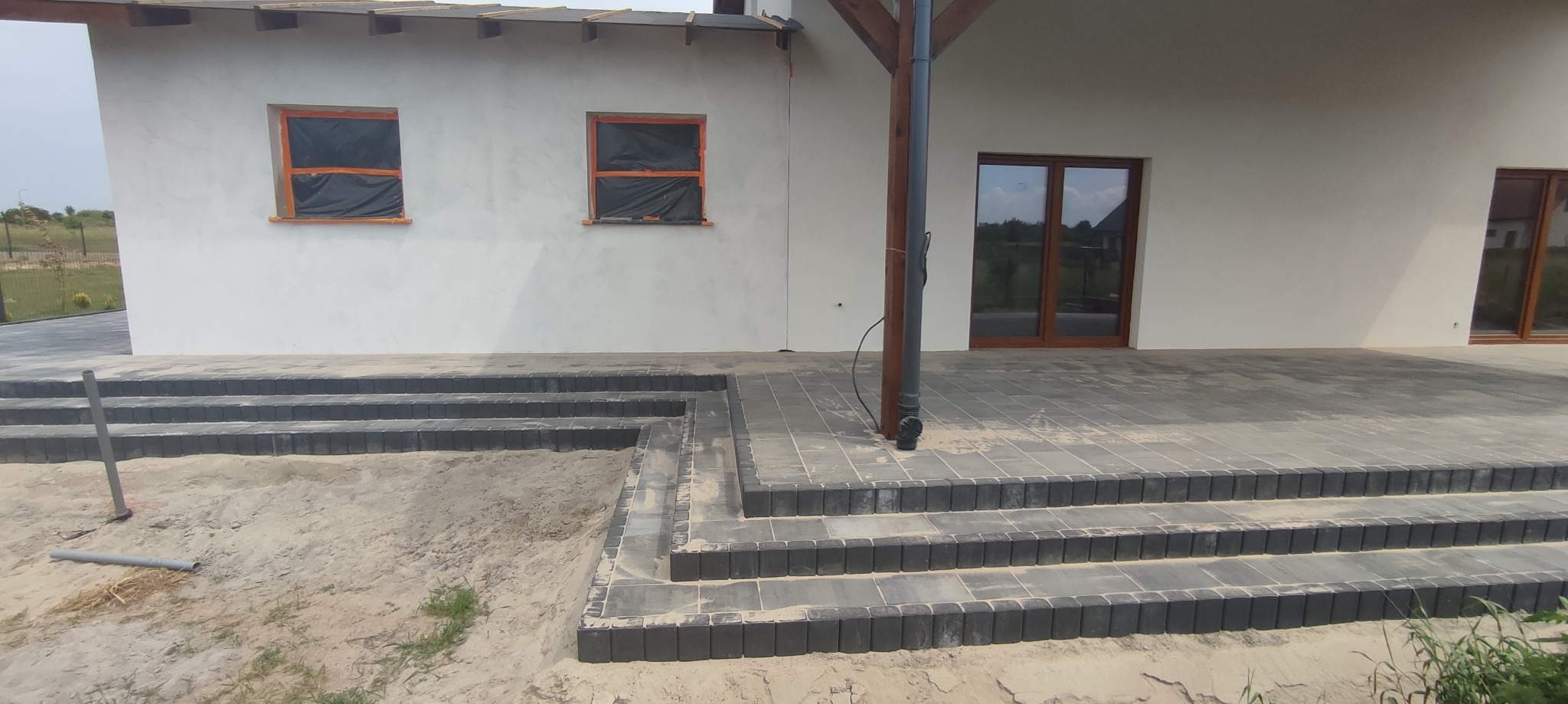 Ułożona kostka brukowa w kolorze grafitowym tworząca schody i taras przy nowym domu z oknami zabezpieczonymi folią, widoczne rury drenażowe na piasku.
