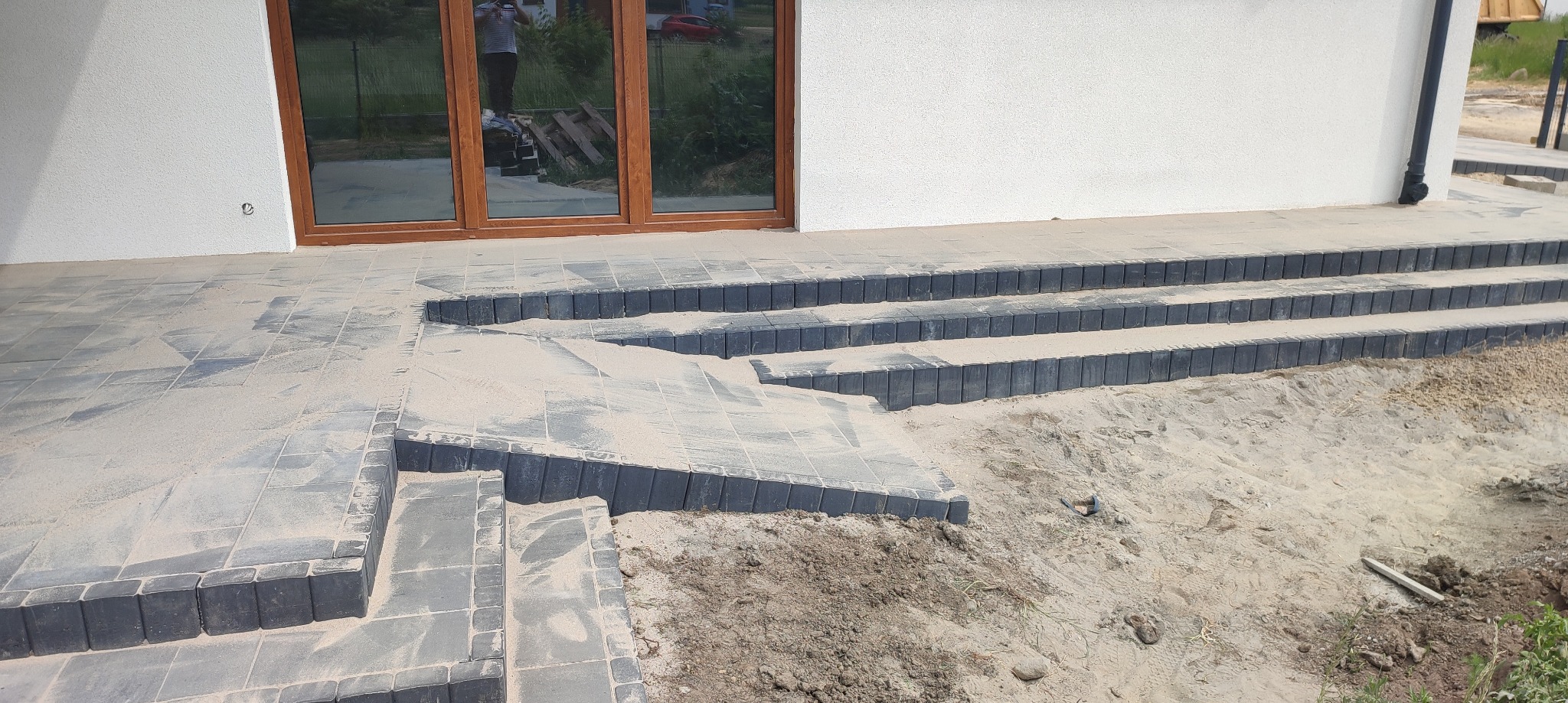 Schody wejściowe do domu wyłożone szarą kostką brukową, częściowo zasypane piaskiem, widoczne okna z odbiciem osoby.