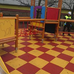 HARTMAN DM SPÓŁKA Z OGRANICZONĄ ODPOWIEDZIALNOŚCIĄ - Kolorowy plac zabaw z motywem wielbłądów, wykonany z drewna i tworzyw sztucznych, z pracownikiem w kasku i kamizelce odblaskowej na tle szachownicy z czerwono-żółtych płyt.