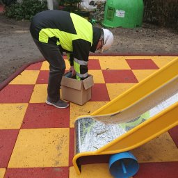 HARTMAN DM SPÓŁKA Z OGRANICZONĄ ODPOWIEDZIALNOŚCIĄ - Pracownik w odblaskowej kurtce i kasku pochyla się nad kartonem obok żółtej zjeżdżalni na placu zabaw z czerwono-żółtą nawierzchnią.