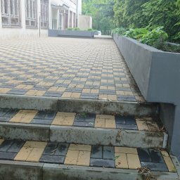 HARTMAN DM SPÓŁKA Z OGRANICZONĄ ODPOWIEDZIALNOŚCIĄ - Schody wejściowe wyłożone kostką brukową w szachownicę z żółto-szarych elementów, prowadzące na plac z identyczną nawierzchnią, obok betonowa donica z zielenią.