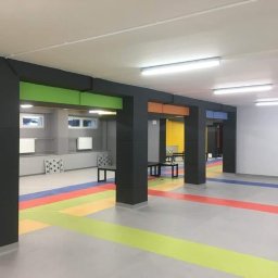 HARTMAN DM SPÓŁKA Z OGRANICZONĄ ODPOWIEDZIALNOŚCIĄ - Wykończone wnętrze z kolorowymi akcentami: szare filary z kolorowymi belkami (żółta, zielona, pomarańczowa, niebieska), podłoga z pasami w kolorach tęczy, białe ściany i sufit z oświetleniem LED.