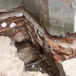 HARTMAN DM SPÓŁKA Z OGRANICZONĄ ODPOWIEDZIALNOŚCIĄ - Odkopane fundamenty ceglane budynku w trakcie prac ziemnych, widoczny fragment ściany z ubytkami i fragment sylwetki mężczyzny w pomarańczowej koszuli w kratę.