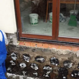 Prace przy fundamentach domu: pracownik w niebieskim kombinezonie podłącza rurę do otworów w ścianie fundamentowej, widoczne ślady po wierceniu i zabezpieczaniu ściany, w tle okno piwniczne.