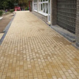 Nowo ułożona, jasnobrązowa kostka brukowa przed budynkiem z białymi oknami i szarą ścianą z luksferów, z widocznymi fragmentami niezabudowanej przestrzeni i schodami z kostki po lewej stronie.