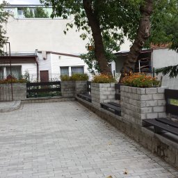 HARTMAN DM Glazurnik Gipsiarz Malarz Sztukator Tapeciarz Szpachlarz Stolarz Dekarz Elektryk Hydraulik Łazienki Kuchnie Remonty Wykończenia Mieszkania - Ustawienie kostki brukowej na podwórku z elementami małej architektury: betonowe donice z pomarańczowymi kwiatami, drewniane ławki i ogrodzenie.