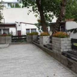 Ustawienie kostki brukowej na podwórku z elementami małej architektury: betonowe donice z pomarańczowymi kwiatami, drewniane ławki i ogrodzenie.