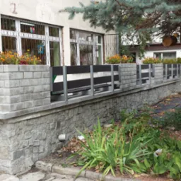 Szary murek z kamienia i cegły z czarnym, poziomym ogrodzeniem i donicami z pomarańczowymi kwiatami przed budynkiem z kratami w oknach.