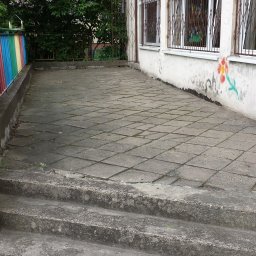 HARTMAN DM Glazurnik Gipsiarz Malarz Sztukator Tapeciarz Szpachlarz Stolarz Dekarz Elektryk Hydraulik Łazienki Kuchnie Remonty Wykończenia Mieszkania - Zewnętrzne schody z betonowych płyt prowadzące na taras z kolorową barierką, ściana budynku z graffiti w tle.