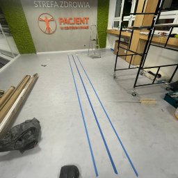 HARTMAN DM SPÓŁKA Z OGRANICZONĄ ODPOWIEDZIALNOŚCIĄ - Przygotowanie podłoża w pomieszczeniu komercyjnym: aplikacja taśmy malarskiej wyznaczającej linie na szarej posadzce, widoczne rusztowanie, drabina, materiały budowlane i narzędzia w tle logo...
