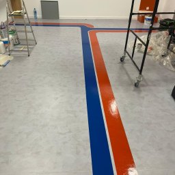 HARTMAN DM SPÓŁKA Z OGRANICZONĄ ODPOWIEDZIALNOŚCIĄ - Świeżo pomalowana podłoga w hali z wyznaczonymi ciągami komunikacyjnymi w kolorach niebieskim i pomarańczowym, widoczne drabiny i materiały malarskie.