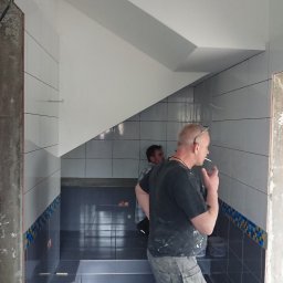 HARTMAN DM SPÓŁKA Z OGRANICZONĄ ODPOWIEDZIALNOŚCIĄ - Wnętrze łazienki w trakcie prac wykończeniowych z widocznymi płytkami w odcieniach szarości i bieli, pracownik z papierosem w ustach oraz częściowo wykończony fragment z dekoracyjnym pasem płytek.