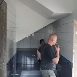 Wnętrze łazienki w trakcie prac wykończeniowych z widocznymi płytkami w odcieniach szarości i bieli, pracownik z papierosem w ustach oraz częściowo wykończony fragment z dekoracyjnym pasem płytek.