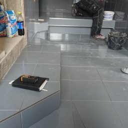 HARTMAN DM SPÓŁKA Z OGRANICZONĄ ODPOWIEDZIALNOŚCIĄ - Układanie szarych płytek w łazience, widoczne narzędzia glazurnicze, torby z zaprawą i pracownicy w trakcie pracy.