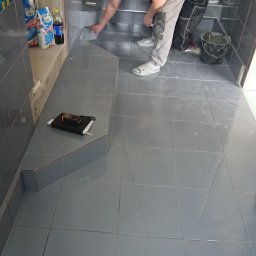 HARTMAN DM SPÓŁKA Z OGRANICZONĄ ODPOWIEDZIALNOŚCIĄ - Mężczyzna w trakcie układania szarych płytek podłogowych w łazience, z widocznym odpływem liniowym i notatnikiem leżącym na podwyższeniu, obok worki z zaprawą.