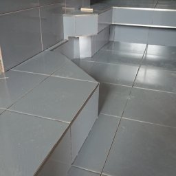 HARTMAN DM SPÓŁKA Z OGRANICZONĄ ODPOWIEDZIALNOŚCIĄ - Szare płytki podłogowe i ścienne z metalowymi listwami wykończeniowymi, fragment nogi w ciemnych spodniach, butelka napoju i worek z zaprawą klejową w rogu.