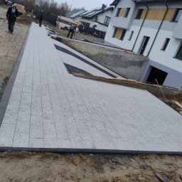DARKABUD - Szeroki, jasnoszary chodnik z kostki granitowej, z czarnymi obrzeżami, prowadzący do nowoczesnego domu z szarą elewacją i ciemnymi oknami, w tle widoczni pracownicy podczas układania kolejnych...