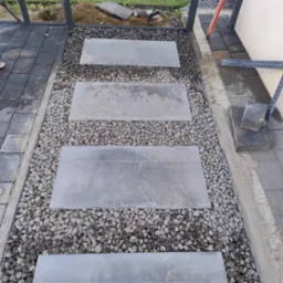 Ścieżka z prostokątnych płyt ułożonych w szarym żwirze, obrzeża z ciemnej kostki brukowej, widoczne elementy metalowej konstrukcji.