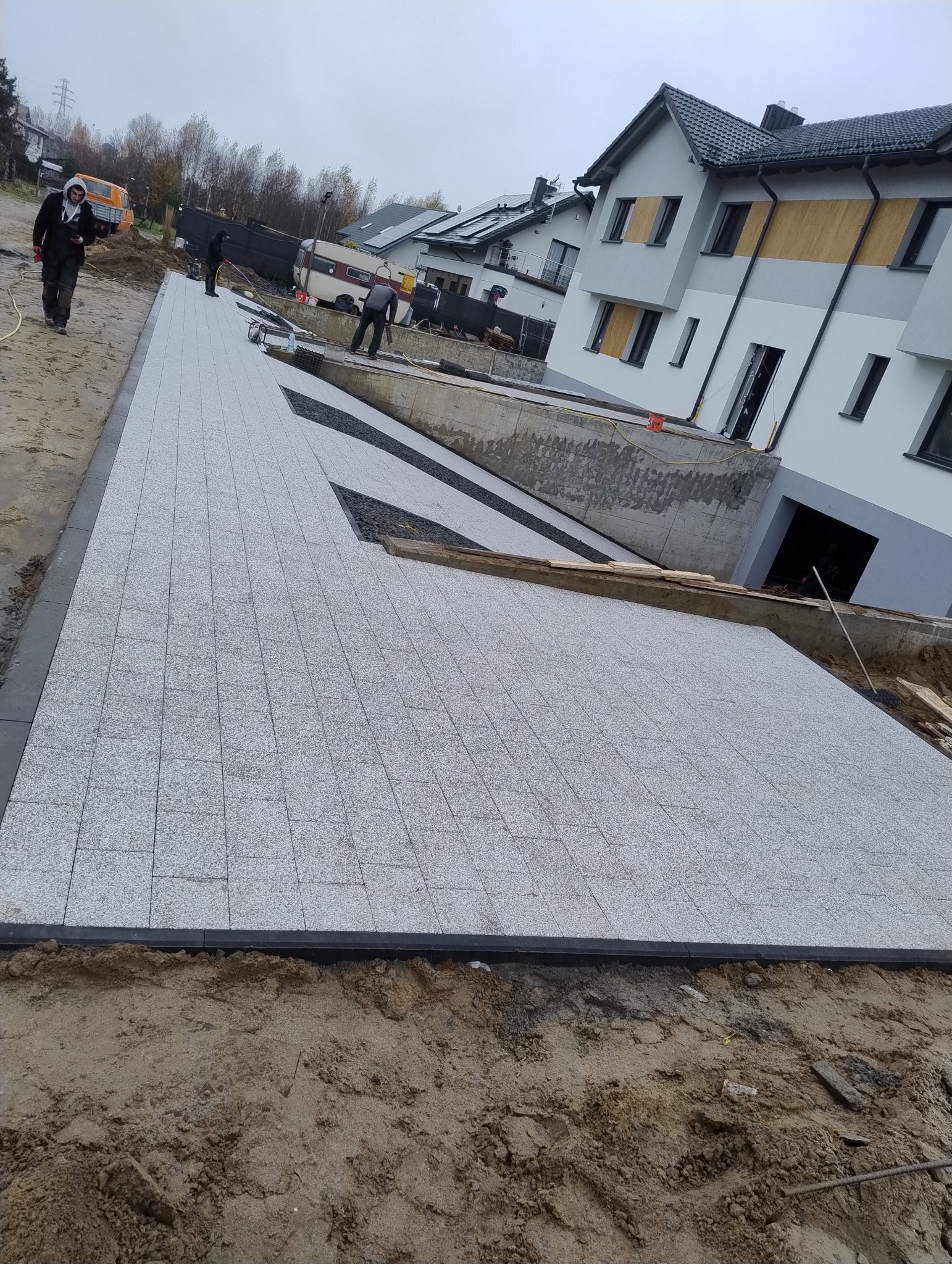 Szeroki, jasnoszary chodnik z kostki granitowej, z czarnymi obrzeżami, prowadzący do nowoczesnego domu z szarą elewacją i ciemnymi oknami, w tle widoczni pracownicy podczas układania kolejnych...