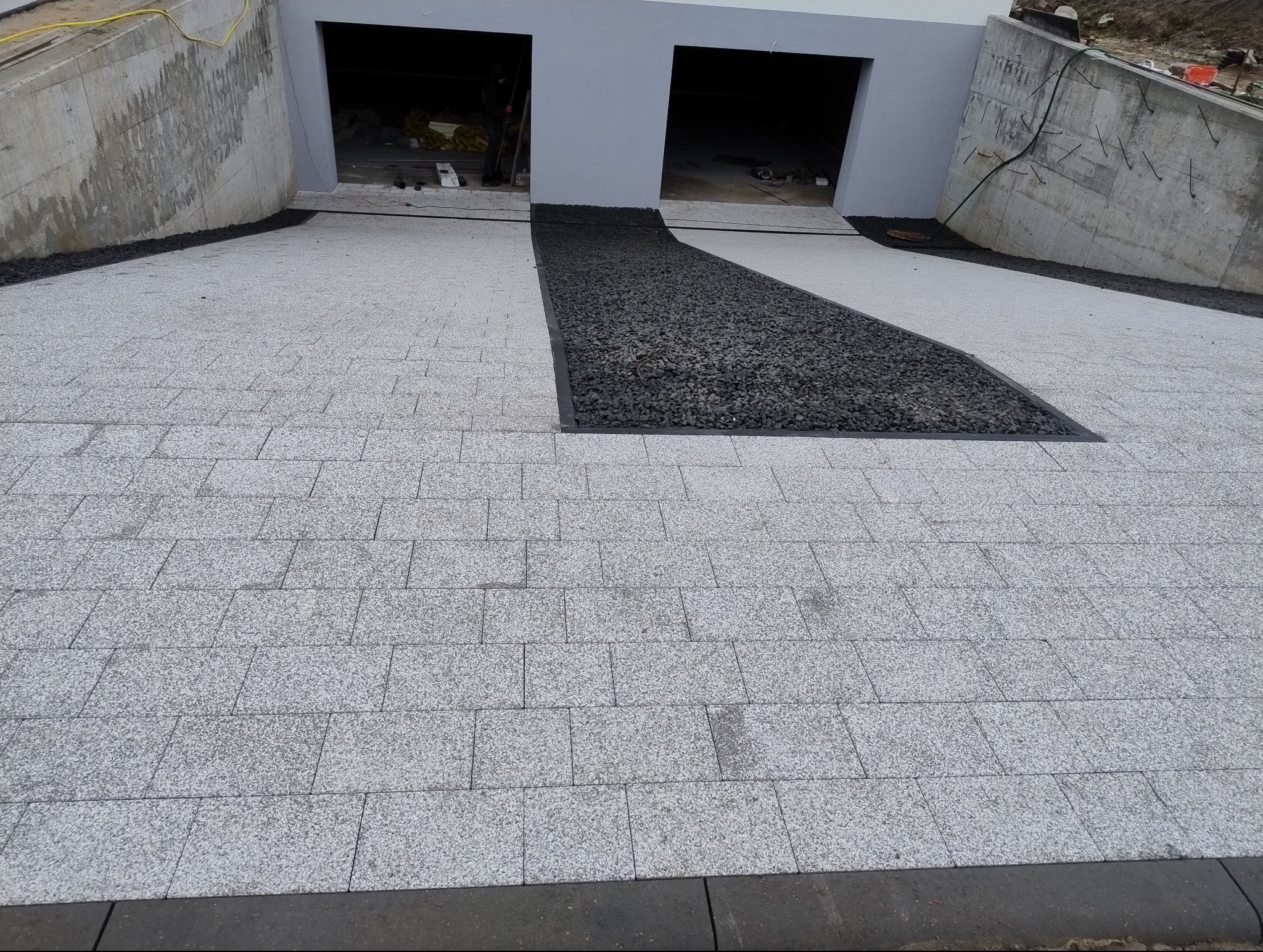 Ułożona jasnoszara kostka granitowa przed wjazdem do garażu, z dekoracyjnym pasem czarnego grysu oddzielonym obrzeżem.