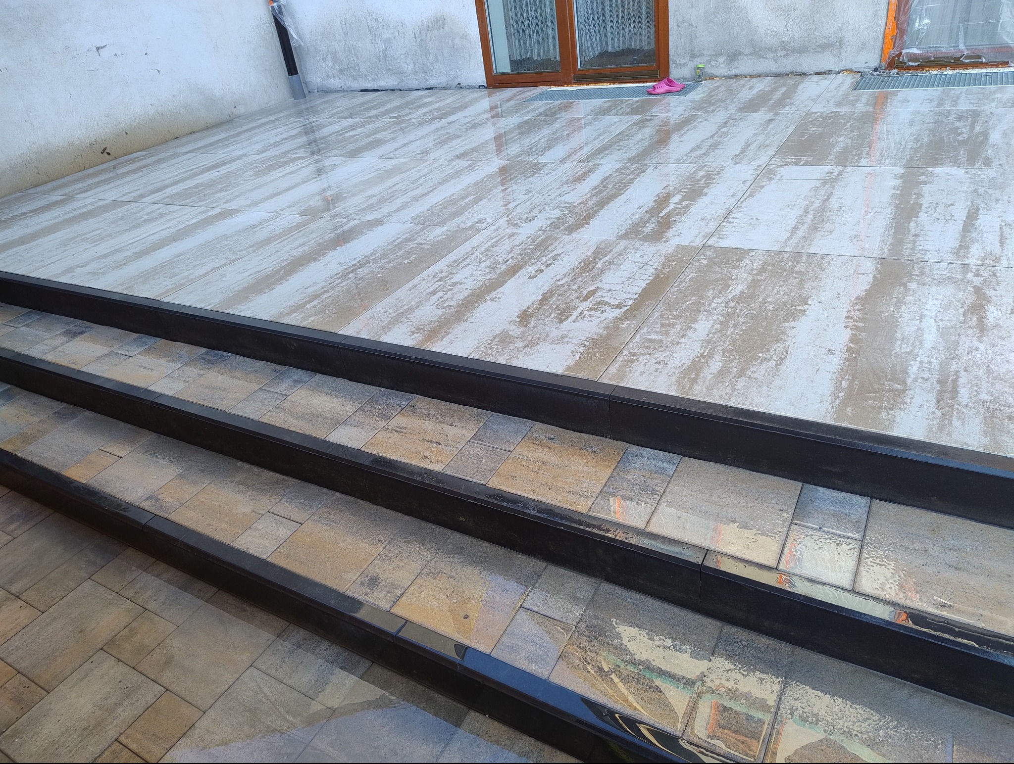 Nowo ułożony, mokry taras z płyt imitujących drewno i schody z ciemną obwódką, widok z dołu na górę, w tle ściana budynku z oknem.