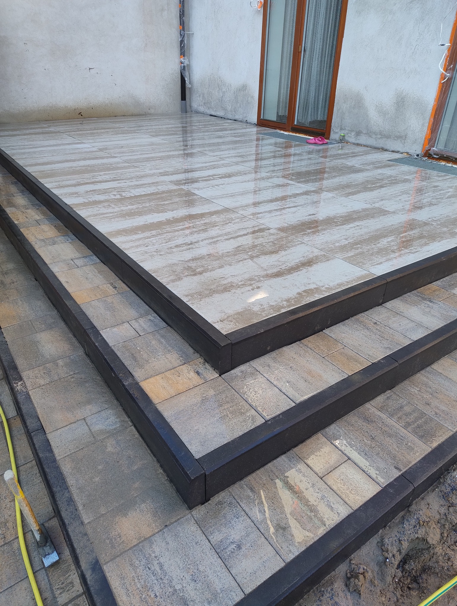 Nowoczesne schody wejściowe wyłożone kostką brukową w odcieniach szarości i beżu, obramowane czarnym obrzeżem, z widocznym fragmentem żółtego węża ogrodowego i narzędzi brukarskich.