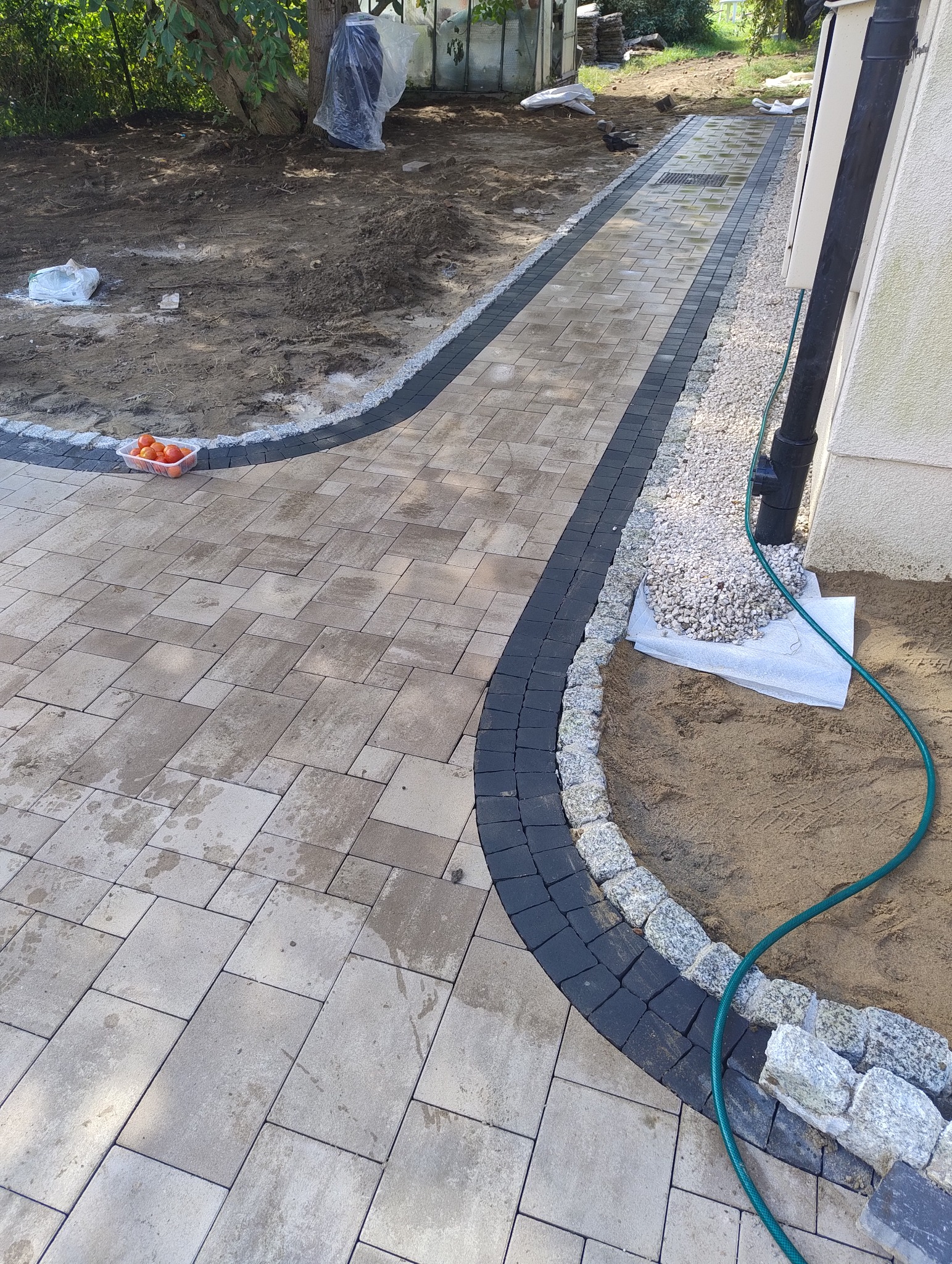 Świeżo ułożony chodnik z kostki brukowej w odcieniach szarości, obramowany ciemniejszą kostką i jasnymi kamieniami, z widoczną warstwą piasku i zielonym wężem ogrodowym, obok ściany budynku.
