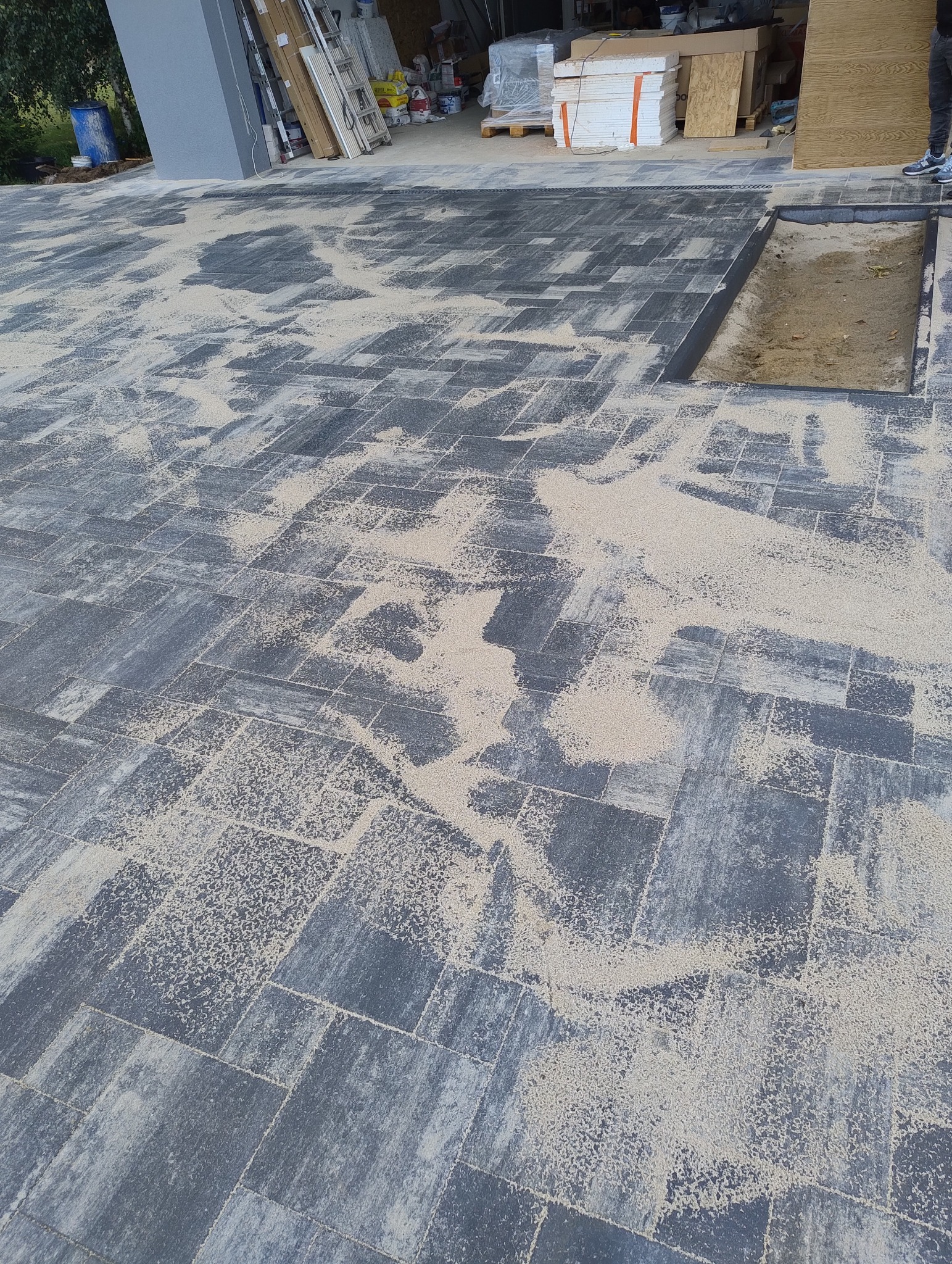 Ułożona szara kostka brukowa z piaskiem w szczelinach, widoczny fragment studzienki rewizyjnej oraz materiały budowlane w tle.