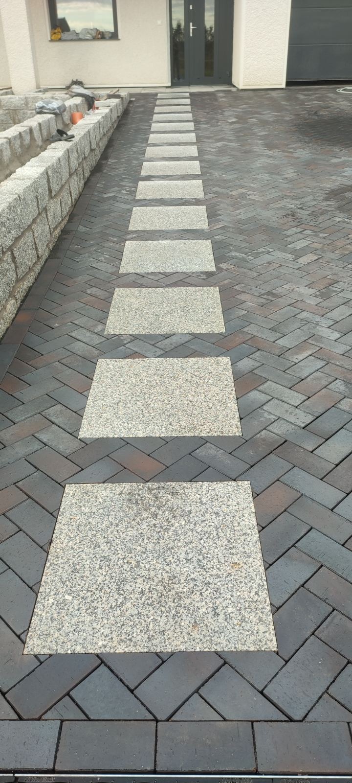Ułożona ścieżka z jasnych, kwadratowych płyt granitowych wkomponowana w ciemnobrązową kostkę brukową ułożoną w jodełkę, prowadząca do drzwi wejściowych domu z szarą elewacją.