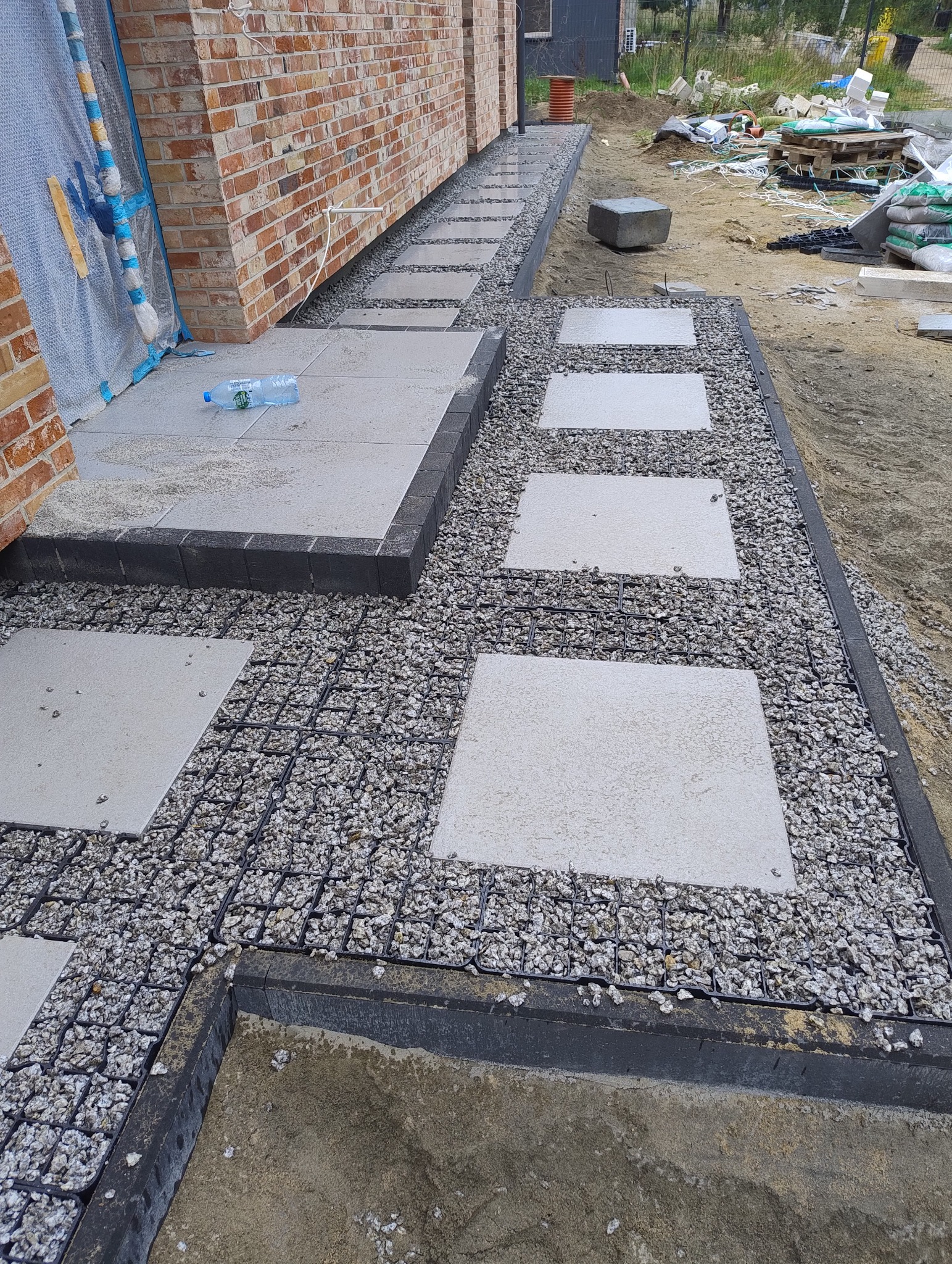 Ścieżka z kwadratowych płyt betonowych ułożonych na siatce wypełnionej drobnym grysem, obrzeża z ciemnoszarej kostki, widok z boku domu z czerwonej cegły, butelka wody na jednej z płyt.