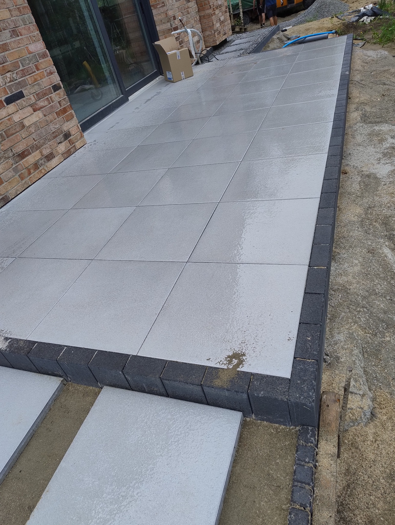 Nowo ułożony taras z jasnych, kwadratowych płyt, obramowany ciemnoszarą kostką, widoczne przygotowane podłoże pod kolejne płyty, w tle fragment ściany z cegły klinkierowej i okno tarasowe.