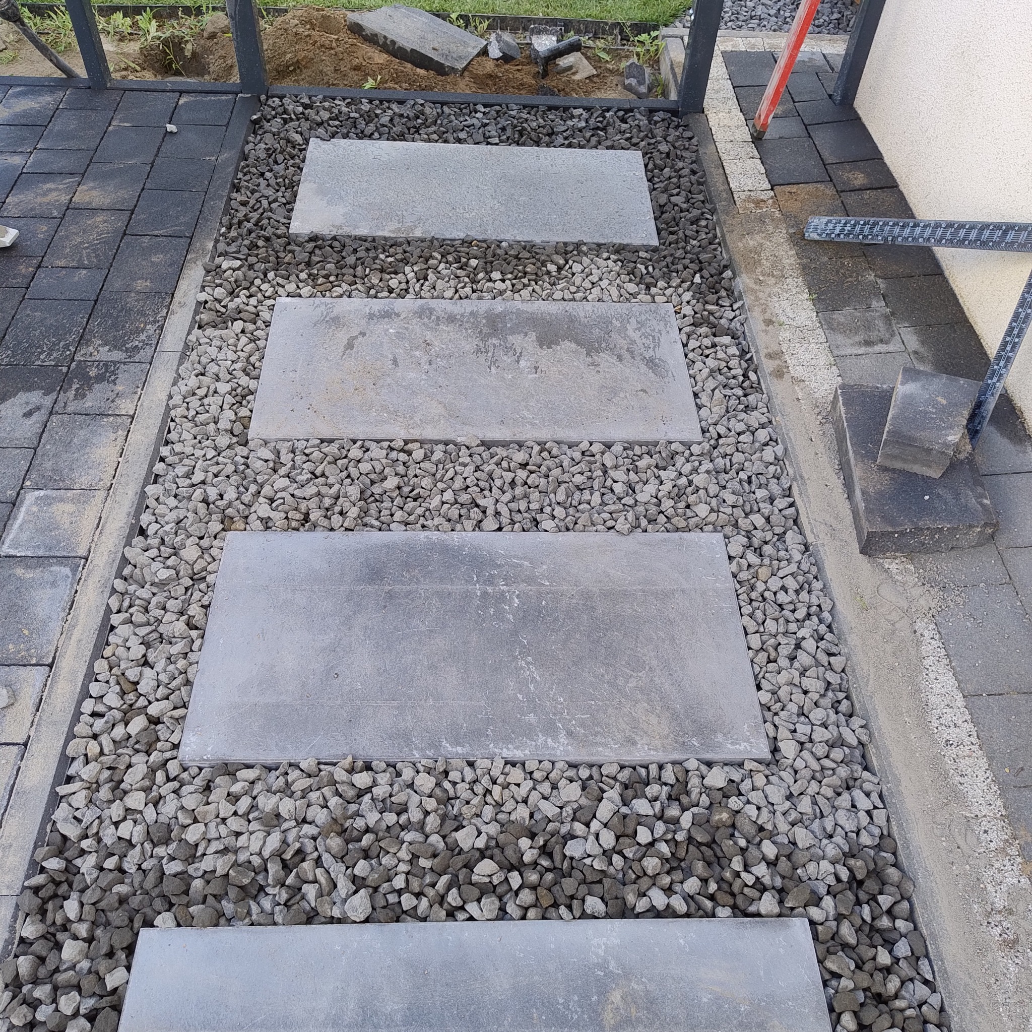 Ścieżka z prostokątnych płyt ułożonych w szarym żwirze, obrzeża z ciemnej kostki brukowej, widoczne elementy metalowej konstrukcji.
