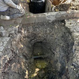 BMP Struzik Piotr - Pracownik w rękawicach klęczy obok wykopu z czarnym zbiornikiem i pomarańczowym wiadrem w środku, widoczne elementy instalacji w wykopie wypełnionym wodą.