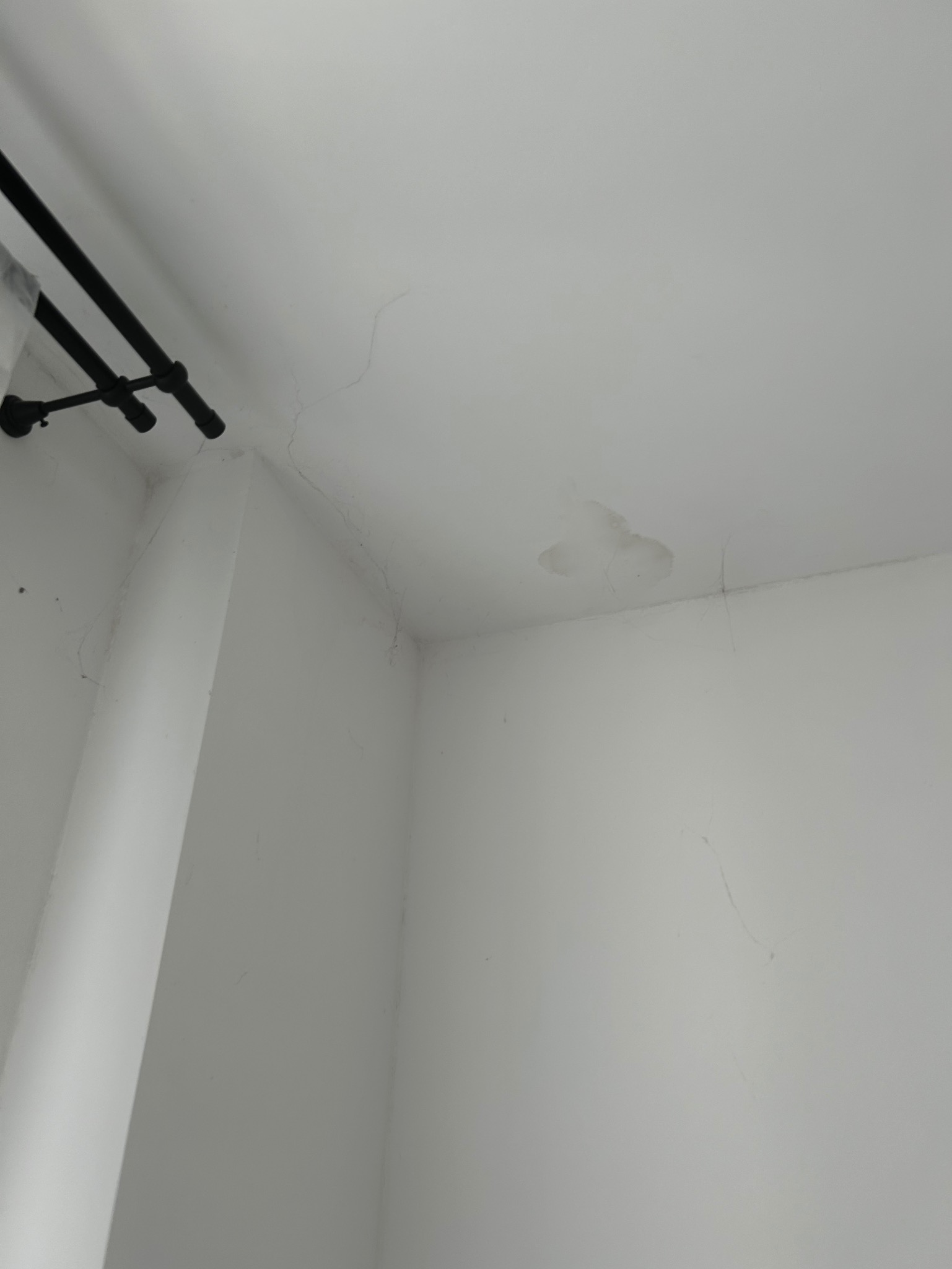 Biały sufit i ściana w rogu pomieszczenia z widocznymi pęknięciami, plamą po zalaniu i czarnym drążkiem karnisza w lewym górnym rogu.