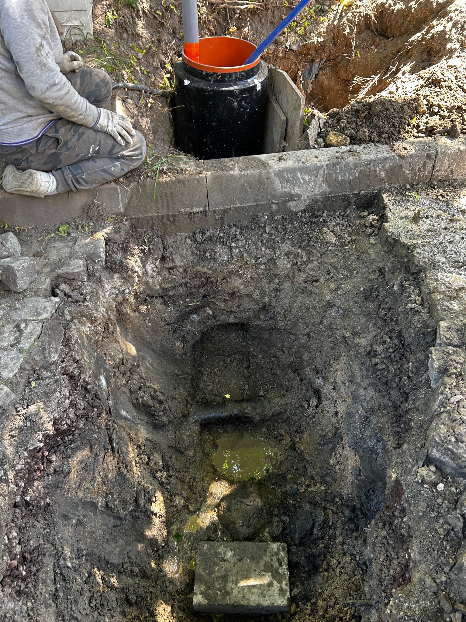 Pracownik w rękawicach klęczy obok wykopu z czarnym zbiornikiem i pomarańczowym wiadrem w środku, widoczne elementy instalacji w wykopie wypełnionym wodą.